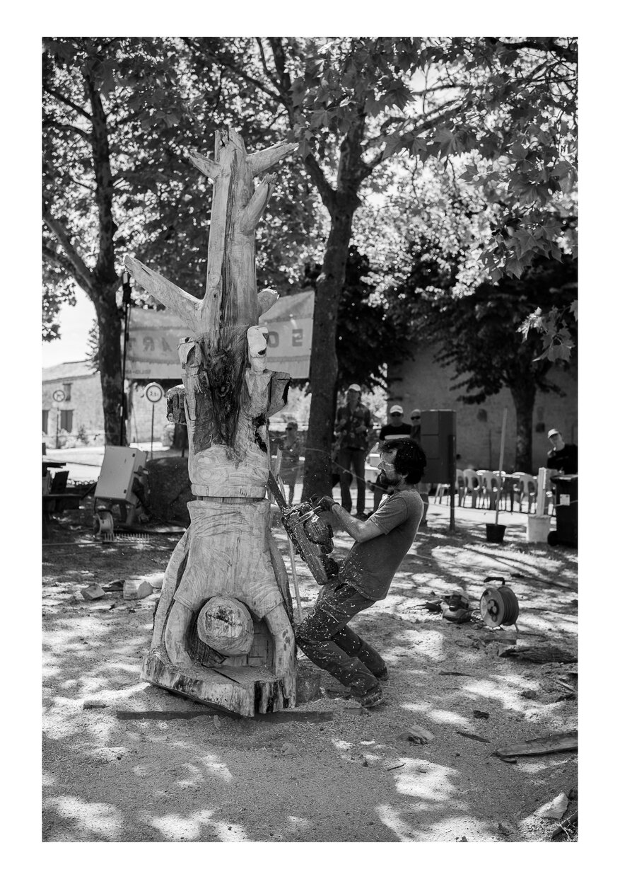 Un homme avec une tronçonneuse façonnant une sculpture en bois (représentant un humain la tête en bas, posé sur ses mains et les pieds en l'air)