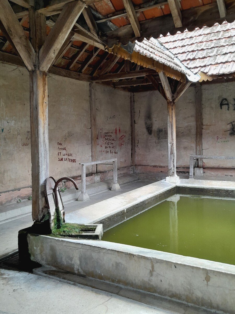 A large old wash basin. The floor around it is covered by a roof, but the basin in the middle is open to the sky. The water looks green and to the left is an old faucet partly covered in moss.