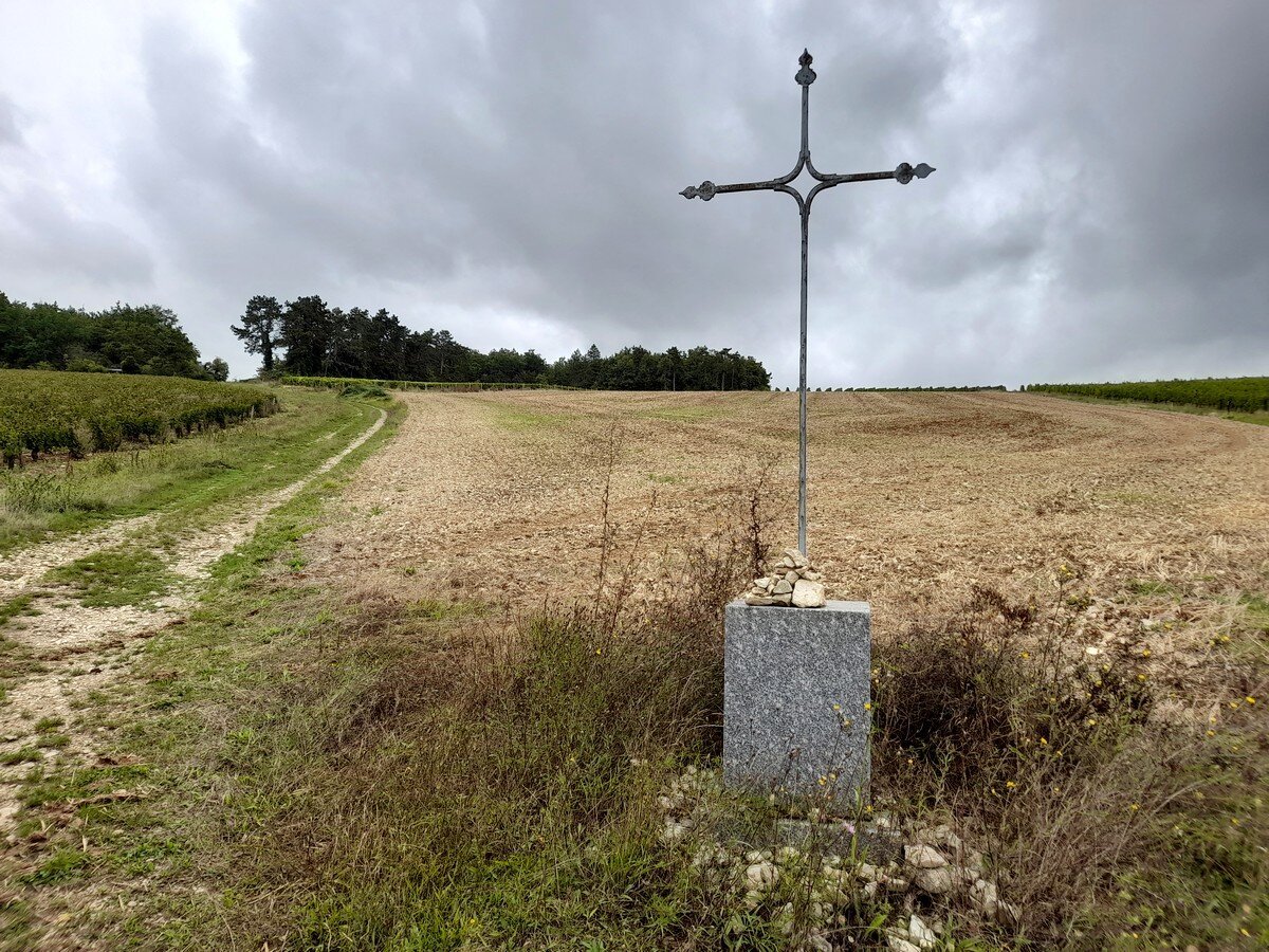 To the right a iron cross in front of a dry field. To the left a path is leading away in the distance towards some trees.