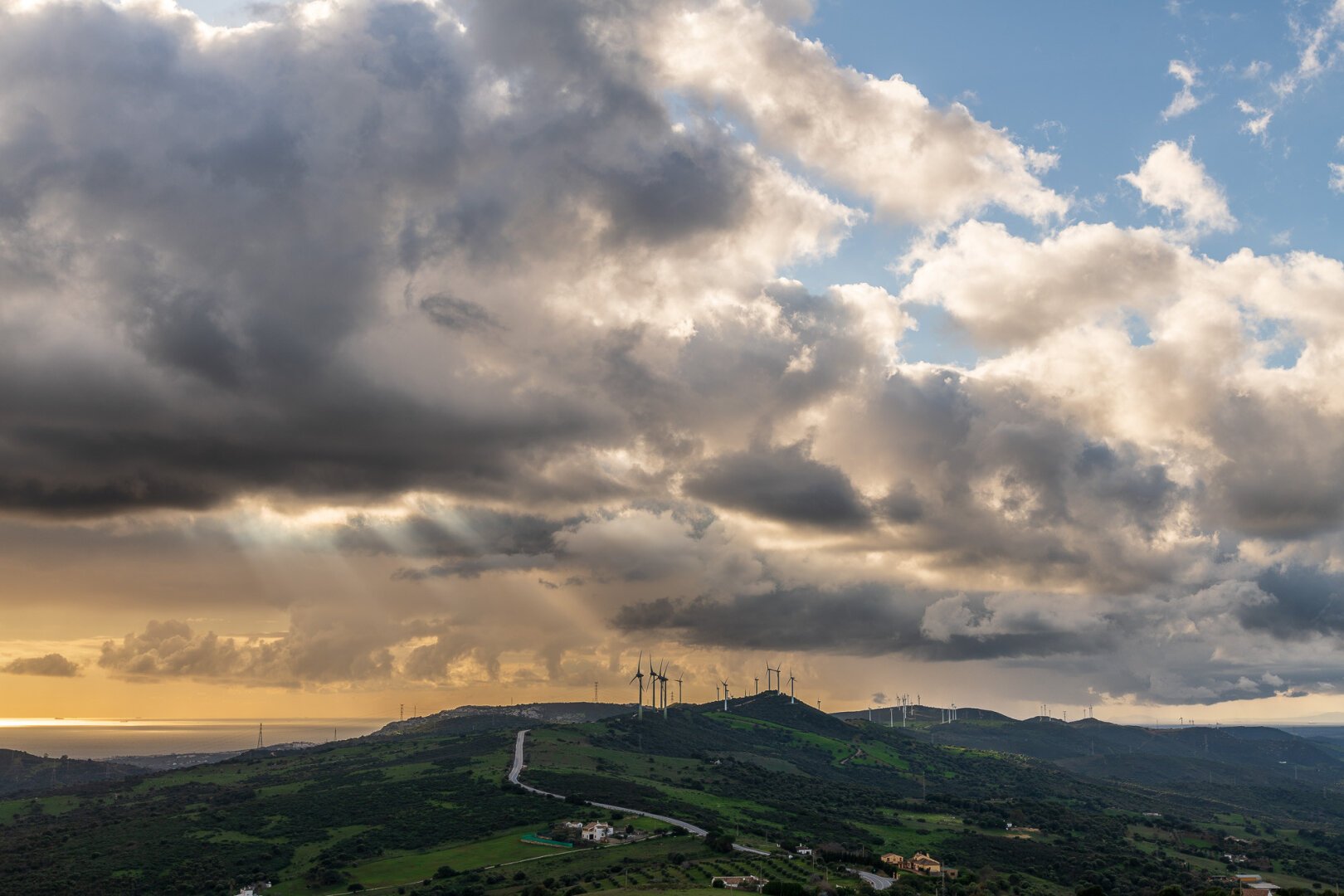 :Una vista panorámica de un paisaje montañoso bajo un cielo dramático. El cielo está lleno de nubes grises y oscuras, con algunos espacios de nubes más claras y brillantes, donde los rayos de sol atraviesan la capa de nubes. En la distancia, se puede ver un mar o océano al atardecer. En las colinas verdes, se observa un parque eólico con varios aerogeneradores. Una carretera sinuosa atraviesa el paisaje. El ambiente general es de tranquilidad y contraste entre la tormenta inminente y la belleza del paisaje.