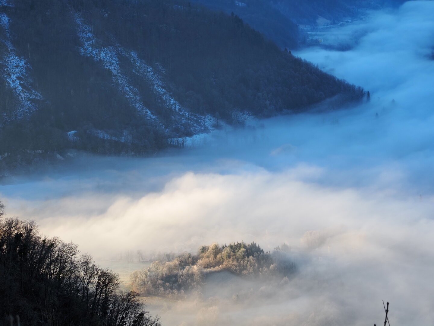 A little hill covered by trees in a valley surrounded by mountains.
The rest of the valley is under a cloud layer. The hill, in the foreground, is illuminated by the sun. The rest of the photo is in the shadow.
