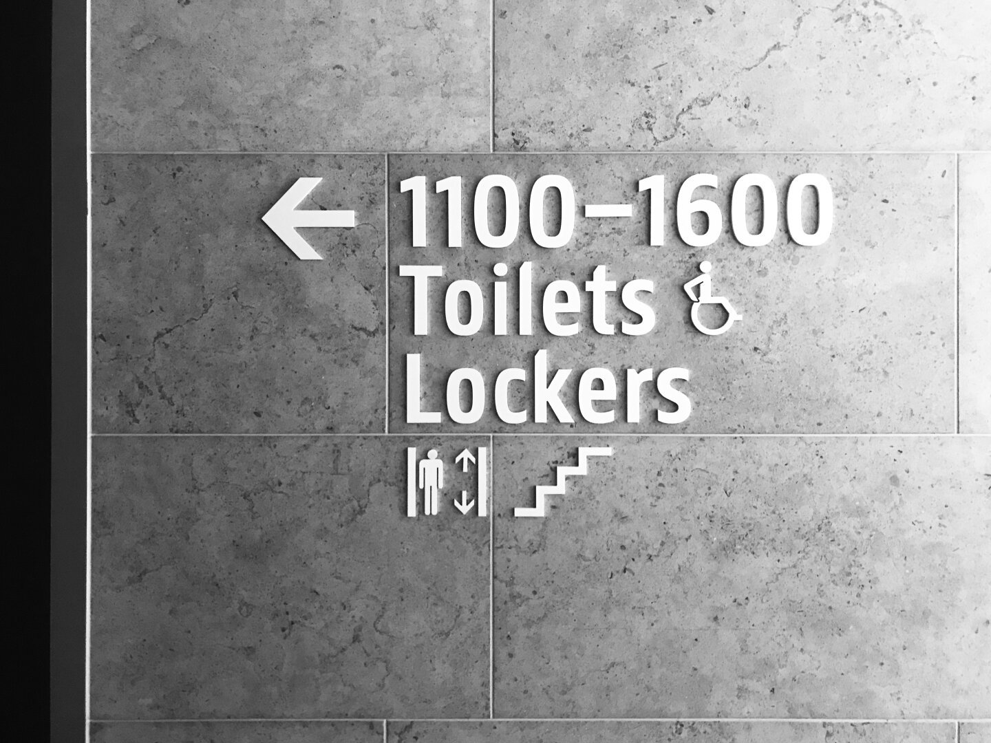 A black and white photograph of a marble wall in a museum that has signage informing visitors that items from the 1100-1600 CE era, the washrooms, lockers and stairs/elevators are all in the same area of the museum.