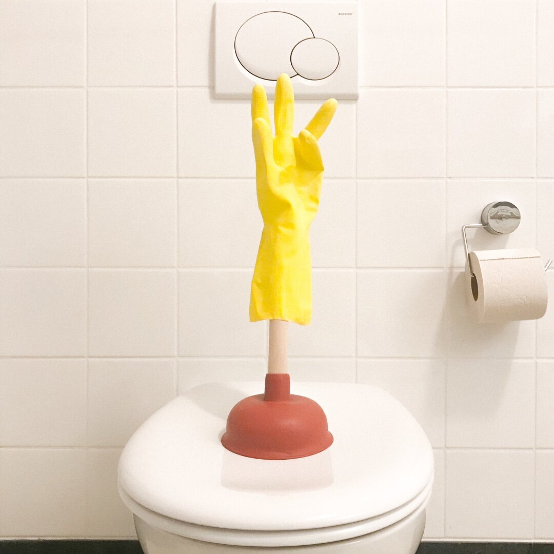 A photograph of a yellow, rubber, cleaning glove that has been impaled on the upright, wooden handle of a toilet plunger — looking a bit like a waving hand, perhaps — and the whole contraption has been placed on a closed toilet seat cover. The surrounding bathroom is a typical all white affair: white tiles; white toilet; white, wall-mounted flush-plate; chrome toilet-paper-holder; unbleached toilet paper roll.