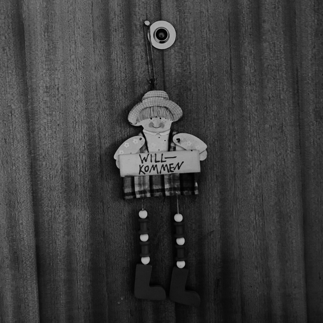 A black and white photograph of a little wooden figure of a boy hanging from a nail on an apartment door. The little boy is holding a sign that says, 