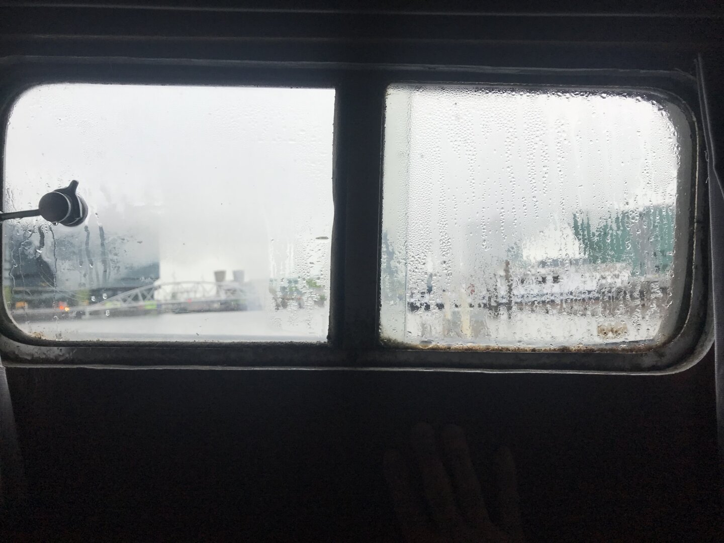 A photograph, taken from inside a former Rhine cargo ship that has now been converted into a small floating hotel. The view is of the Oosterdok — an inner harbour area in Amsterdam, just north of the old city centre — taken through a small, condensation covered, cabin window. In the slightly-out-of-focus, droplet-and-fog-obscured harbour, some buildings and a medium-sized pedestrian bridge are partially visible but hard to identify. The water is calm but the weather is grey and uninspiring. It all makes for a fairly drab scene but it evokes a kind of appropriate melancholy for a cold morning on the water.