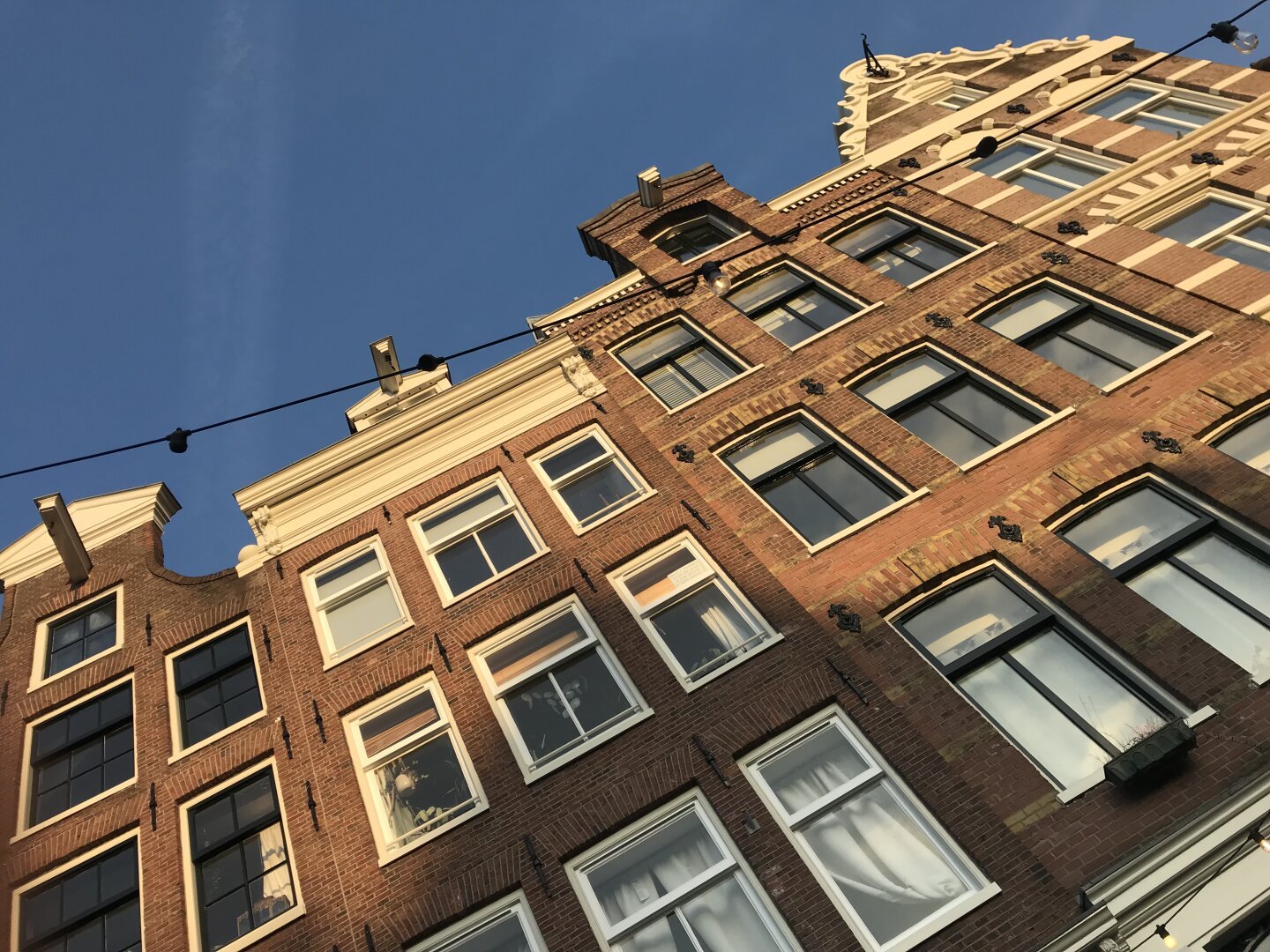 A photograph, taken at a very oblique, upwards angle, of the front facades of some older buildings in Amsterdam. It is a nice, late spring evening with a clear blue sky and the evening sun lights the facades with a warm light. As is typical of historic buildings in the old centre of Amsterdam, the facades feature artistic brickwork; large, wood framed windows; gabled roofs and, at the very tops, hooks for ropes that allow heavy objects to be lifted into, and out of, the thin, tall buildings.