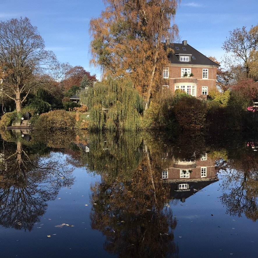 A photograph taken on a path along the Alster Canal in Hamburg, looking across the water at the backyards of two houses that line the canal. The calm, mirror-like waters perfectly reflect the blue sky and the many trees and bushes growing along the canal's edge. On the right side of the photo is a large, brick house with white framed windows typical of older, nicer homes in northern Germany. It is also reflected in the canal. The house on the left side is barely visible behind a large weeping willow tree but their small wooden dock can be seen at the water's edge.