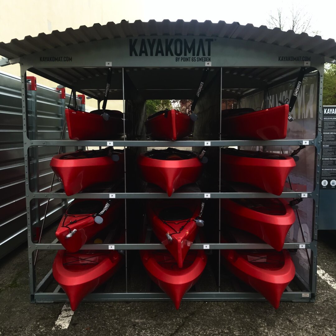 A photograph of the front ends of twelve, red kayaks stacked in a metal-frame structure. They are stacked three to a row and four rows high. Each kayak has a black chain-lock attached to it. The metal structure, most likely aluminum, has an arched, corrugated metal roof. The structure is an automated kayak dispenser located in a small parking lot — and occupies basically one parking space — next to a (not seen in this photo) canal in Hamburg, Germany.