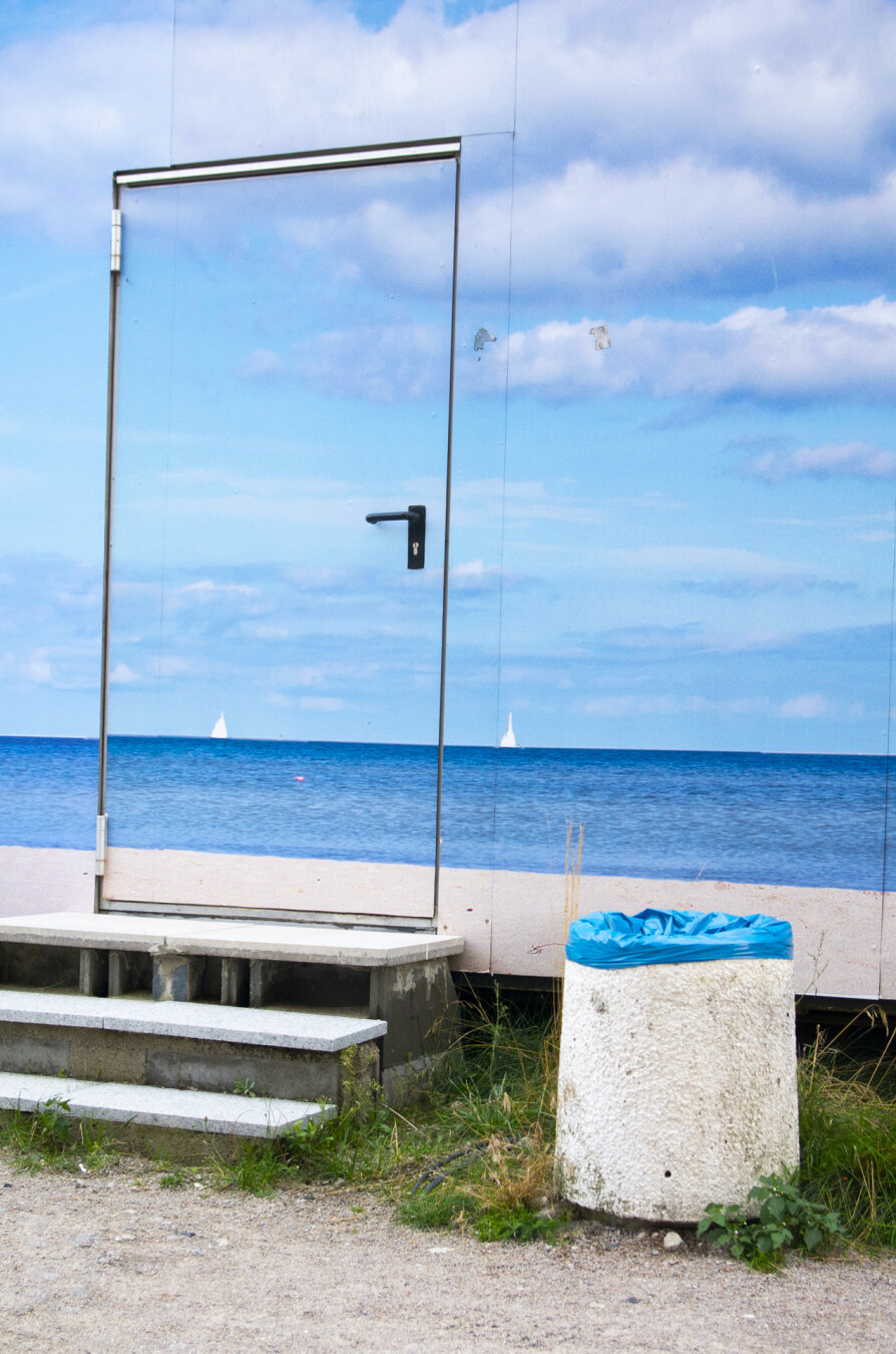 Color photograph, portrait orientation. A construction trailer covered with images of the ocean view, sand, the sea, and the sky, ranging from very blue to light blue; toward the left-center of the photo is a doorframe and three steps; on the right is a light-colored trash can with a blue bag, with sand and green plants in front of it.

farbfotografie, hochformat. ein baucontainer, beklebt mit meerblick, sand, meer und himmel, sehr blau bis hellblau; halblinks auf dem foto ein türrahmen, drei treppenstufen, rechts ein heller mülleimer mit blauer tüte, davor sand und grüne pflanzen.