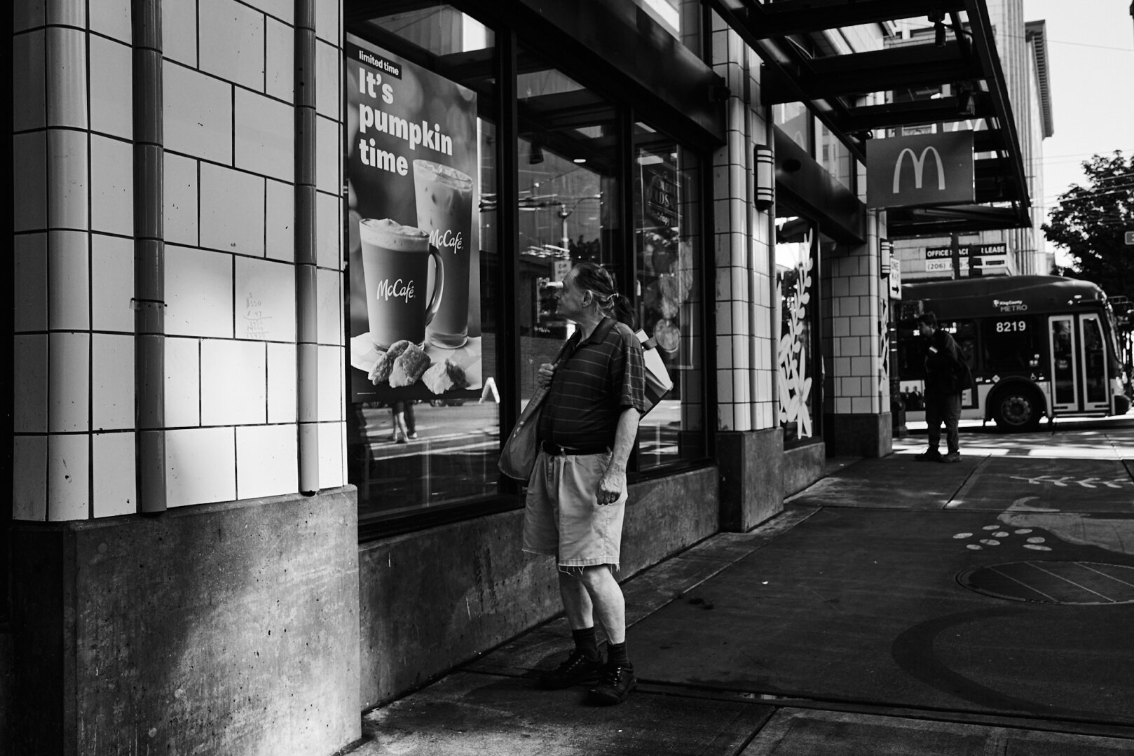 Black and white photo of a man standing on a sidewalk in the Central Business District of Seattle, looking at a window display advertising McCafe's pumpkin-themed drinks. There is a bus in the background.