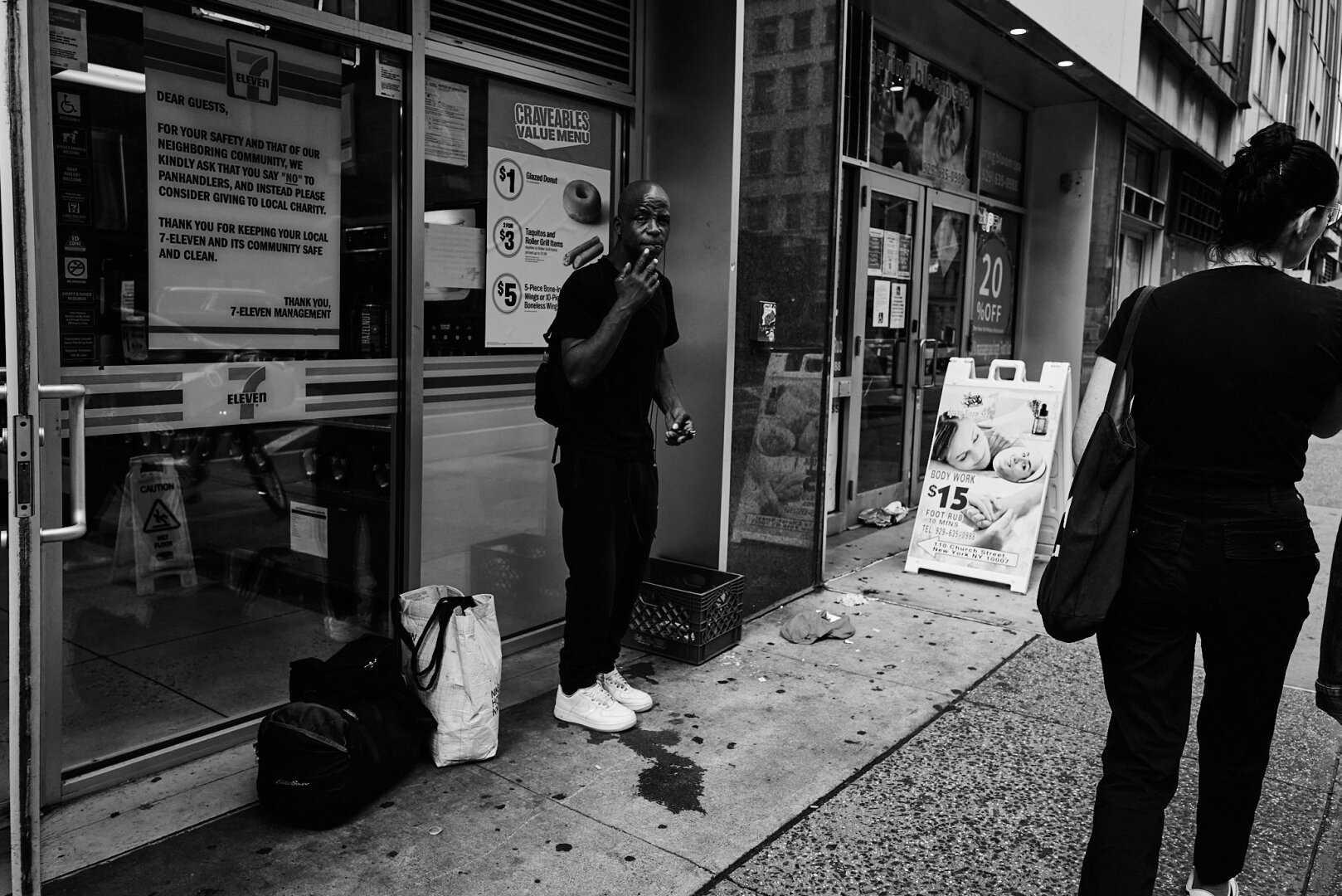 A black and white image of a person standing outside a 7-Eleven in Tribeca, New York City, holding a cigarette. There are bags on the ground nearby, and a sign in the window addresses panhandling.