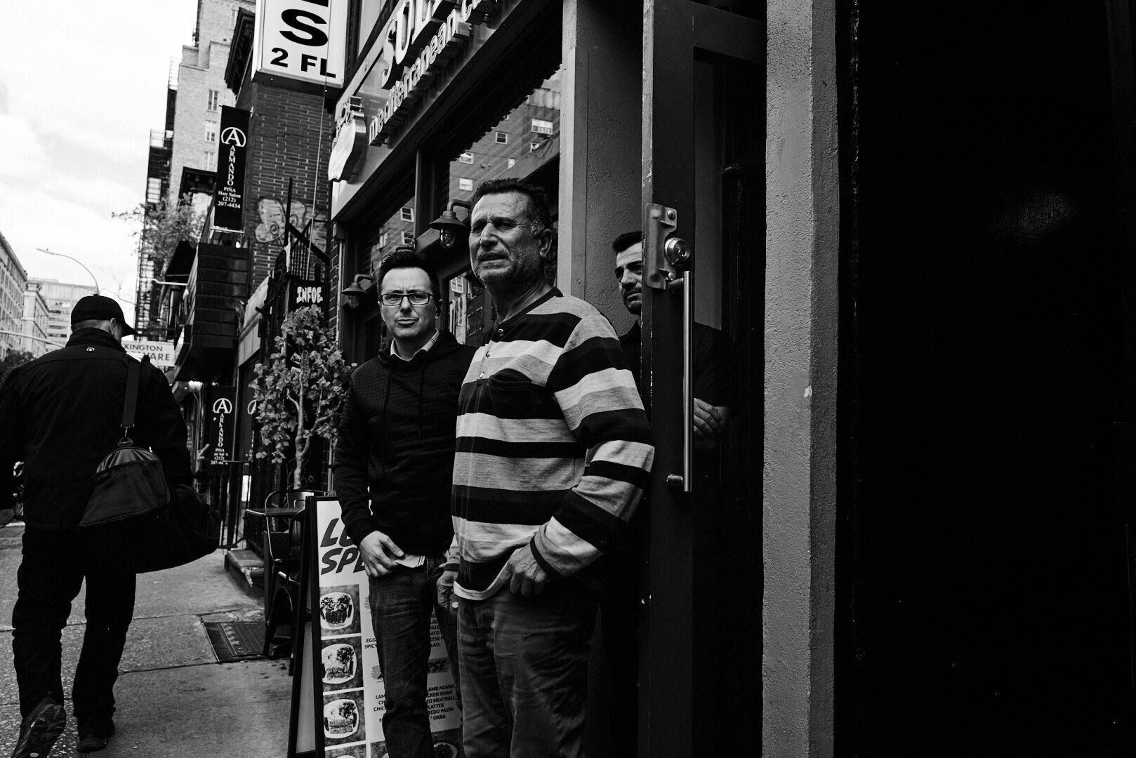 Black and white street photo of three men standing near an open doorway in Manhattan. One man is wearing a striped shirt, and another wears glasses. A passerby in a black jacket walks along the sidewalk.