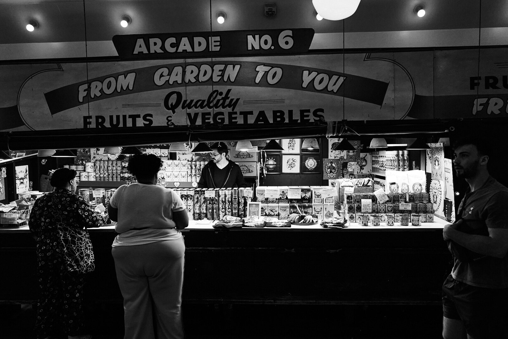 Black and white photo of a market stall in Belltown, Seattle, showcasing a vendor behind a counter filled with various packaged goods. Two customers stand in front, browsing the items lit by overhead lights.
