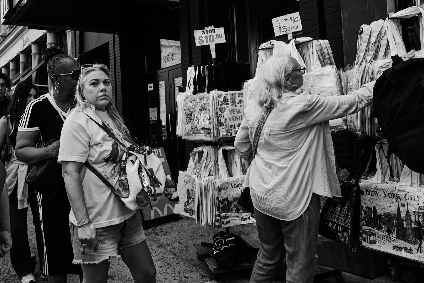 A black and white photo of three people standing in front of a street stall selling New York City-themed tote bags. Two women are browsing the selection, while one woman looks directly at the camera with a serious expression. Signs advertise pricing for the bags.