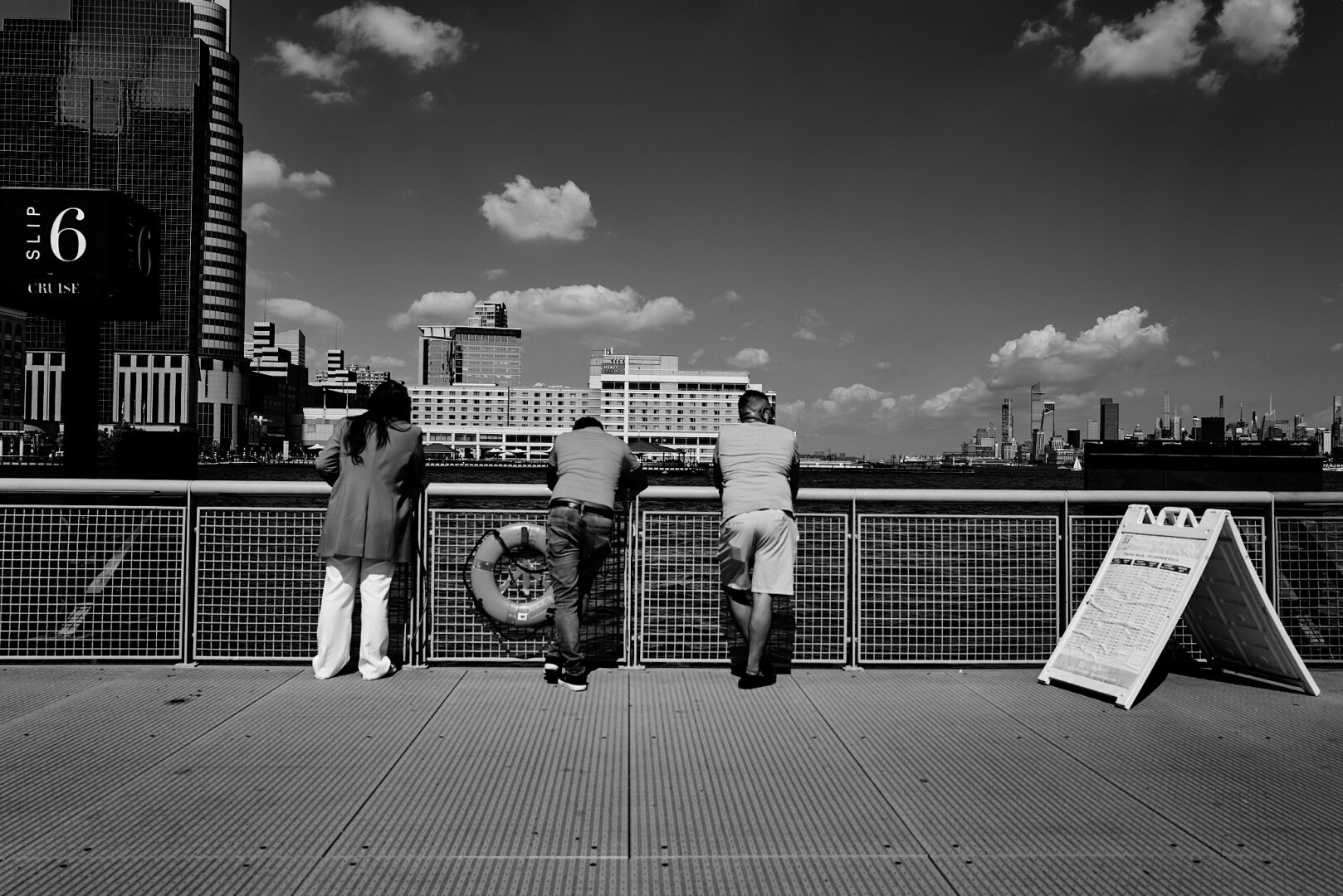 Three people leaning against a metal railing overlooking a waterfront in Jersey City, with buildings visible in the background. The scene is captured in black and white under a clear sky.