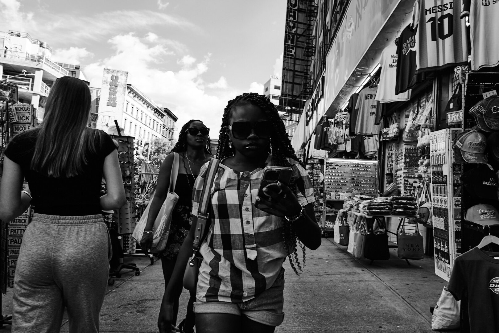 Black and white image of a bustling city sidewalk with people walking past shops. A woman in the foreground is wearing sunglasses and looking at her phone, while other pedestrians move in different directions.