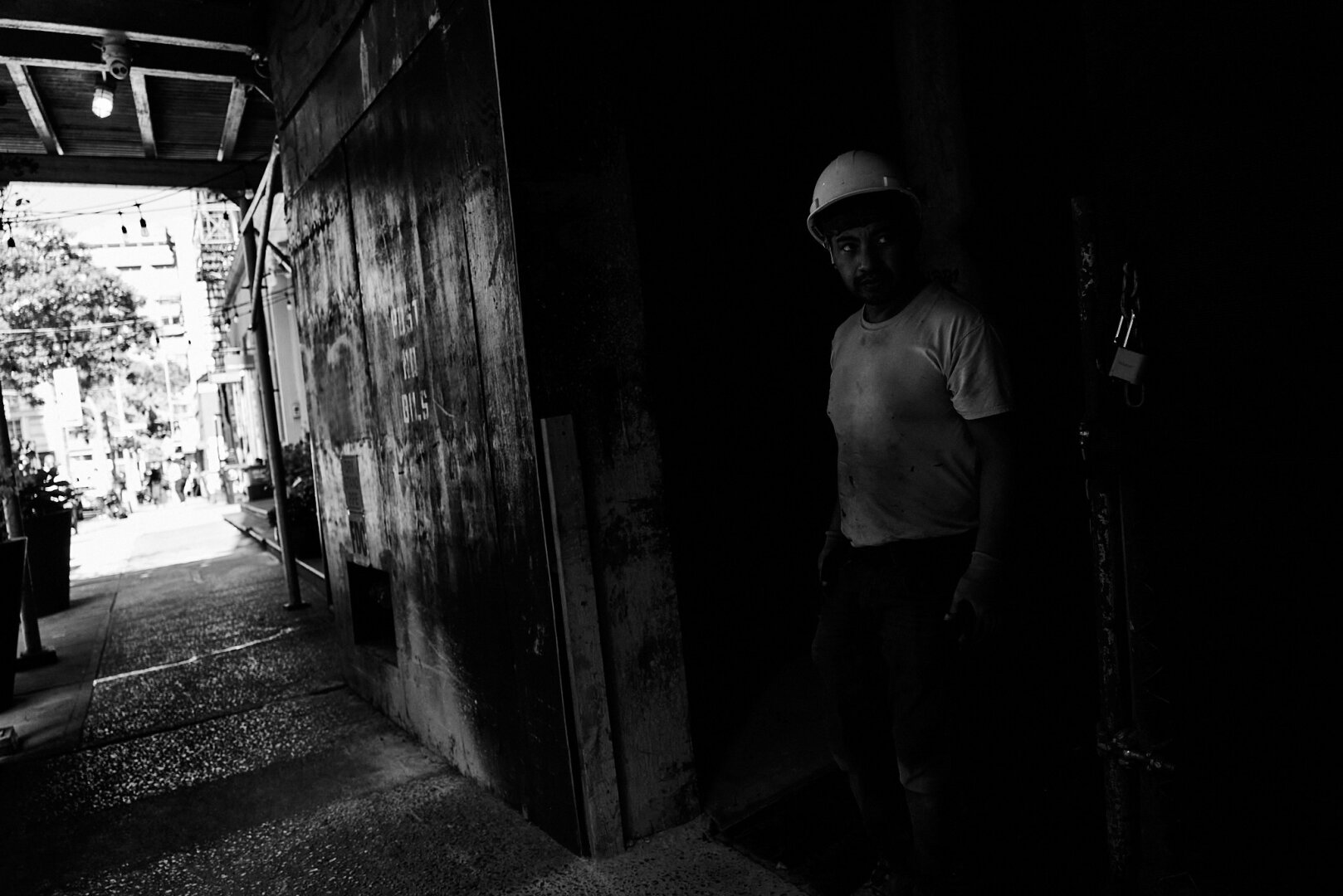 A man wearing a hard hat stands partially in the shadow of a dark construction site entrance, with a bright urban scene visible in the background.