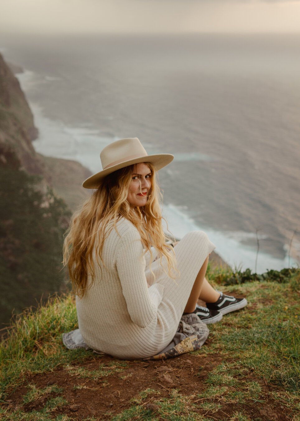 A woman in light-colored clothing and hat, sits on a hillside, with the sea and cliffs in the background. She looks directly into the lens.