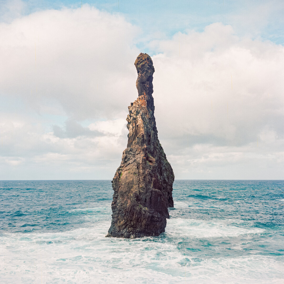 Film photo of a tall solitary volcanic rock spire rising dramatically from the ocean waves off the coast of Madeira, Portugal, under a cloudy sky, with white foam crashing at its base.