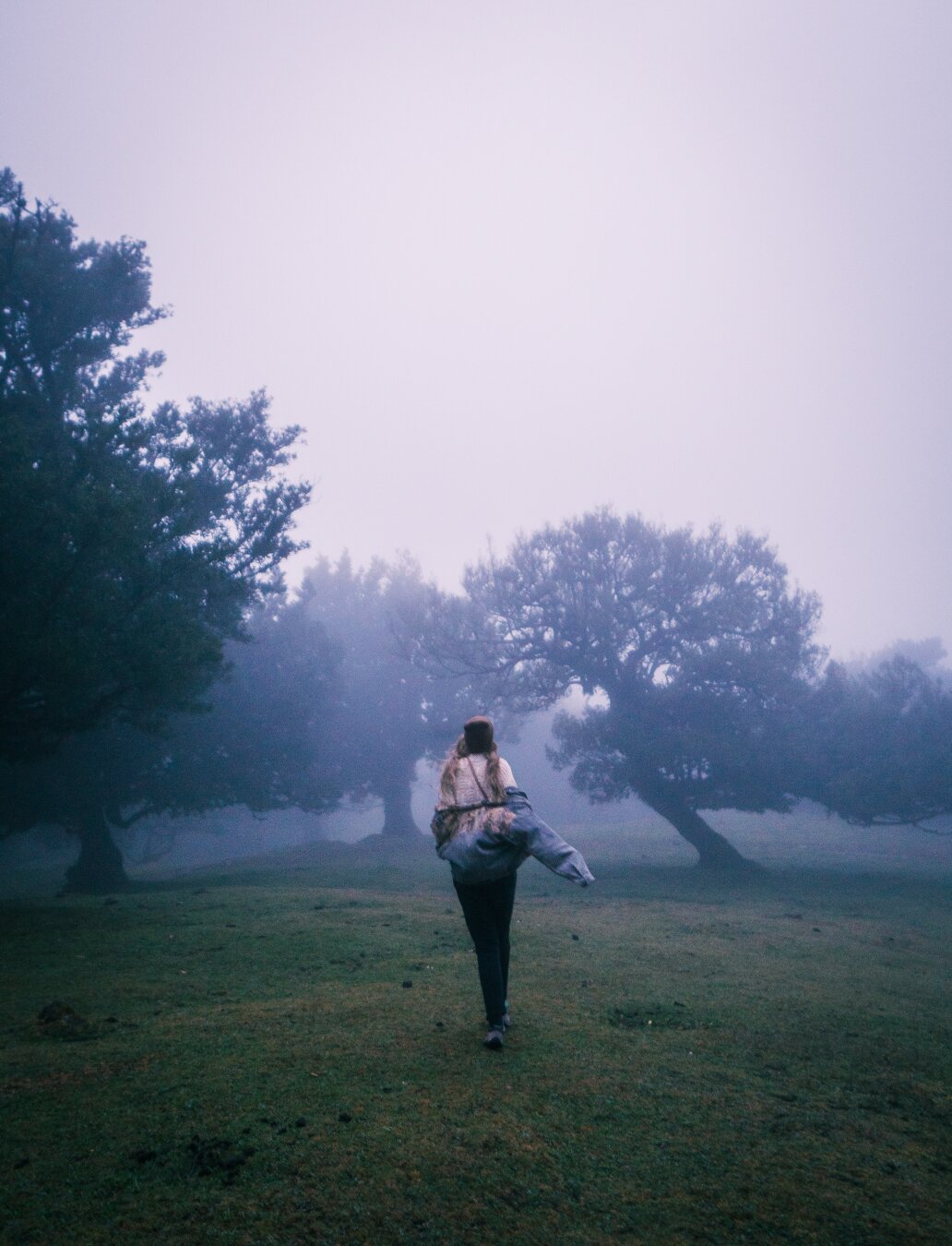 A person walking playfully with one arm raised through a foggy meadow near Faial, Madeira, Portugal, silhouetted trees fading into thick mist in the background.