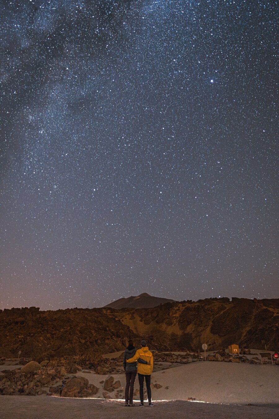 Two people standing together on a rocky volcanic landscape at night, gazing up at a breathtaking star-filled sky with the Milky Way visible overhead. One person wears a bright yellow jacket. The rugged silhouette of Mount Teide rises in the background, suggesting a location high in Teide National Park, Tenerife.