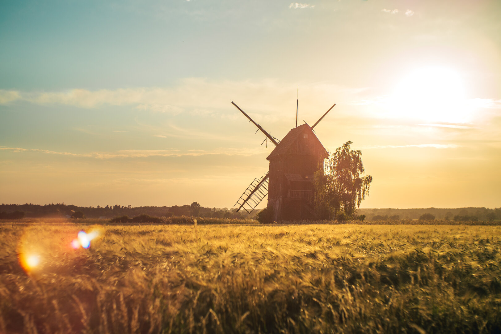A rustic wooden windmill silhouetted against a warm golden sunset, standing beside a tree at the edge of a wheat field, with lens flares scattered across the frame.