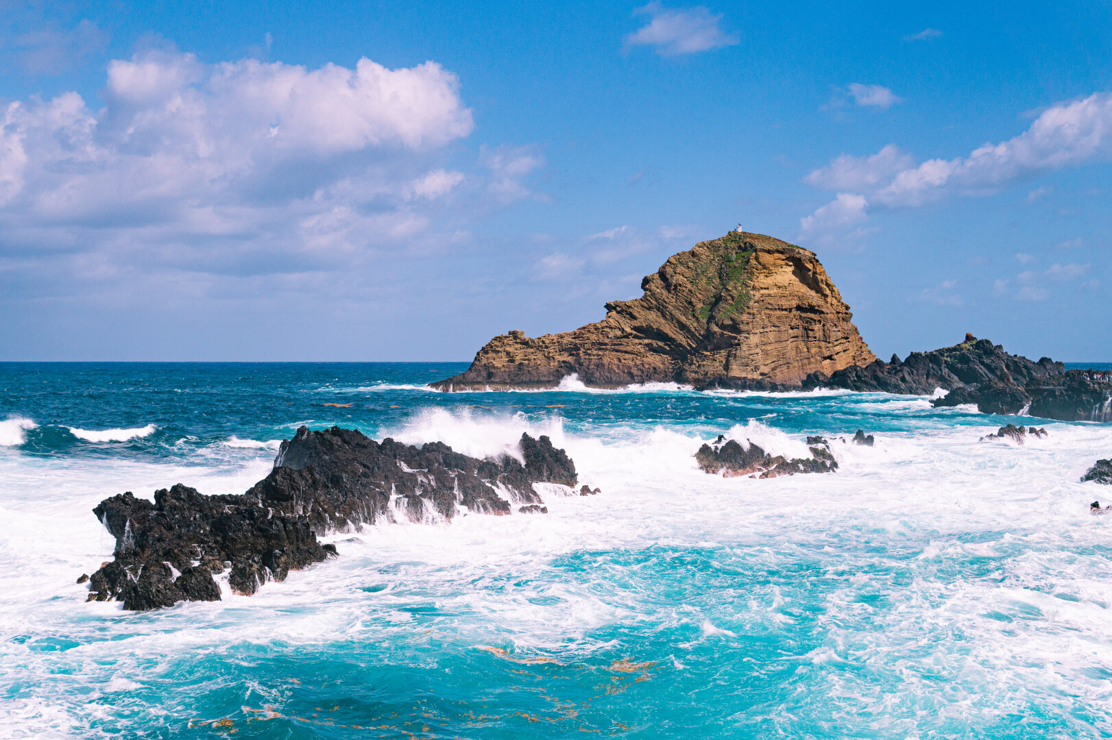 Large layered rock formation off the coast of Porto Moniz, Madeira, Portugal, with dark volcanic rocks in the foreground and turquoise waves crashing against them under a bright blue sky.