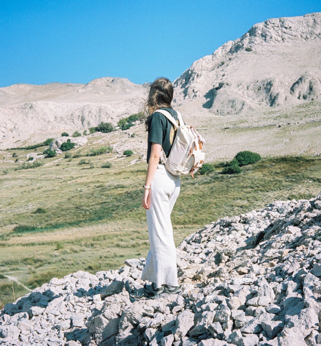 Woman with a backpack standing with her back to the lens, behind her a view of the stone hills