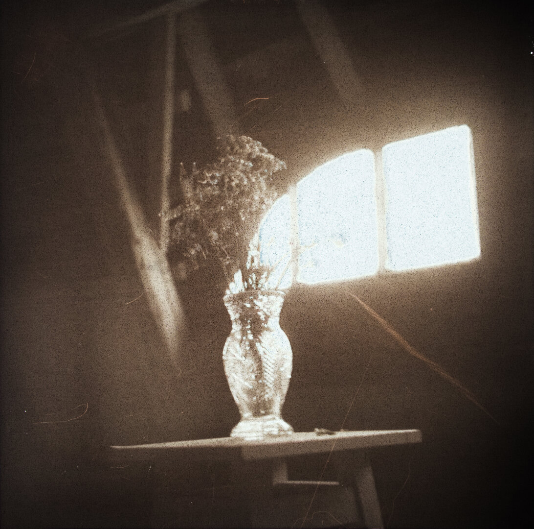 A crystal vase with a bouquet of dried flowers on a white stool in the attic, with wooden beams and a window visible in the background.