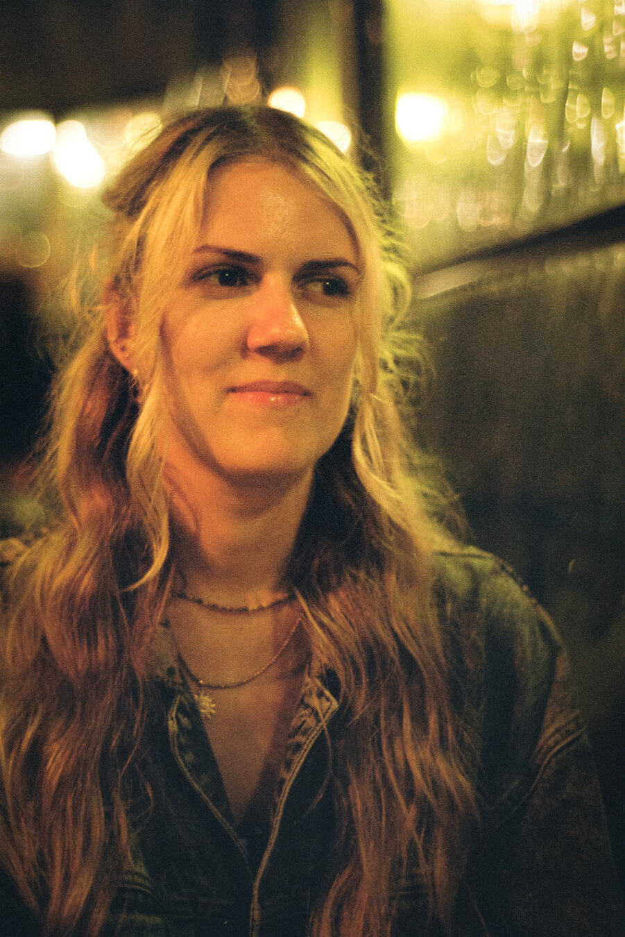 A portrait of a woman looking to the side, she has long, slightly curly blonde hair, she is wearing a grey denim jacket, blurred windows can be seen in the background.