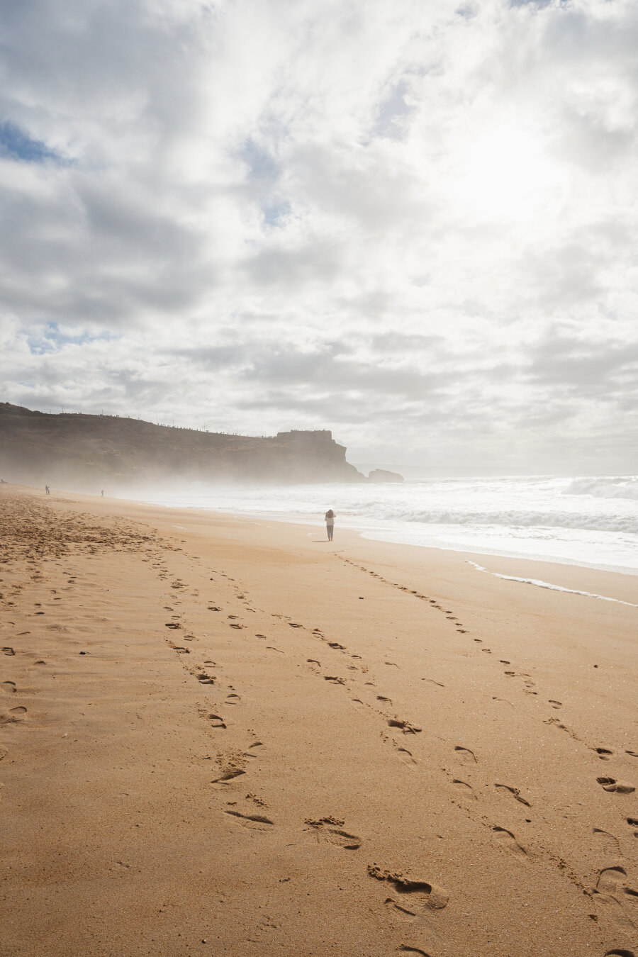 A beach with lots of mist, a woman stands in the middle. In the background is an overcast sky and cliffs.
