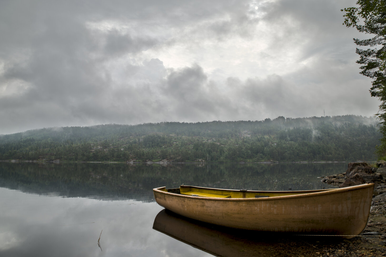 bild im querformat. im vordergrund liegt am ufer ein gelb-oranges ruderboot, dahinter ein ruhiger, sich spiegelglatter see an dessen anderen ufern ein hügel mit bäumen wächst, in dem nebel hängt. die sonne ist hinter dichten wolken zu erahnen