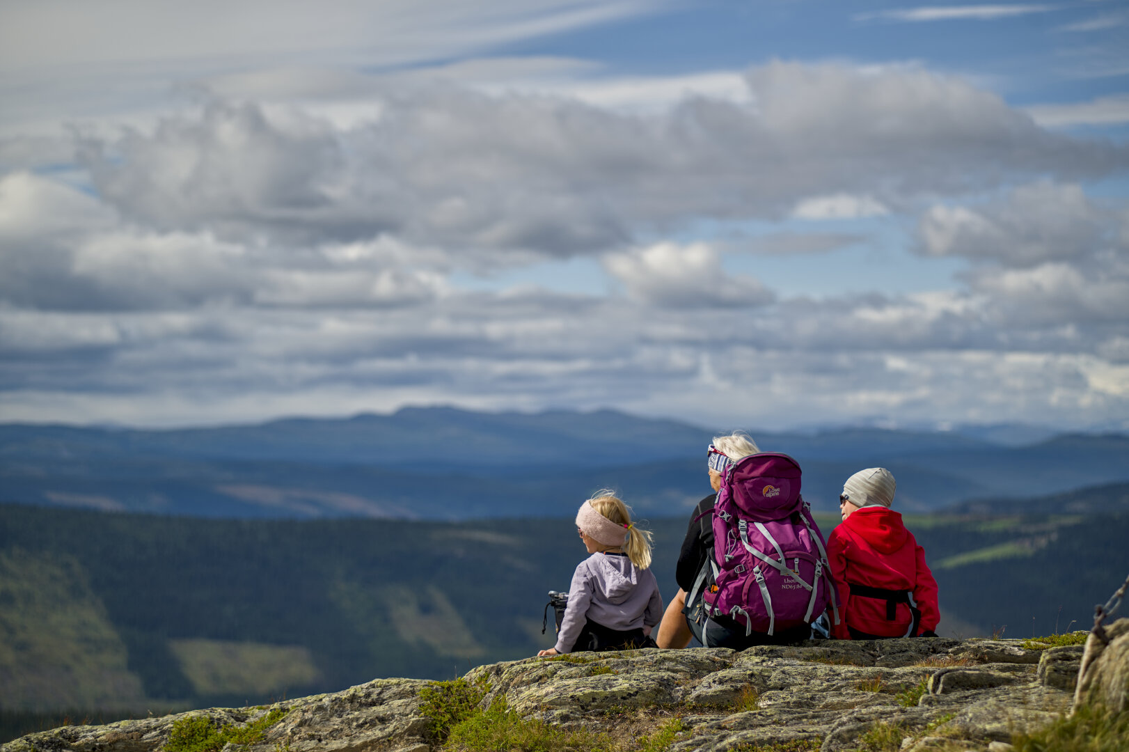 bild im querformat auf einer berg"spitze"... 
im rechten bilddrittel sitze eine frau, links und rechts von ihr je ein kind, alle gucken nach links richtung soone.
im hintergrund wolken vor leicht blauen himmel und weitere berketten