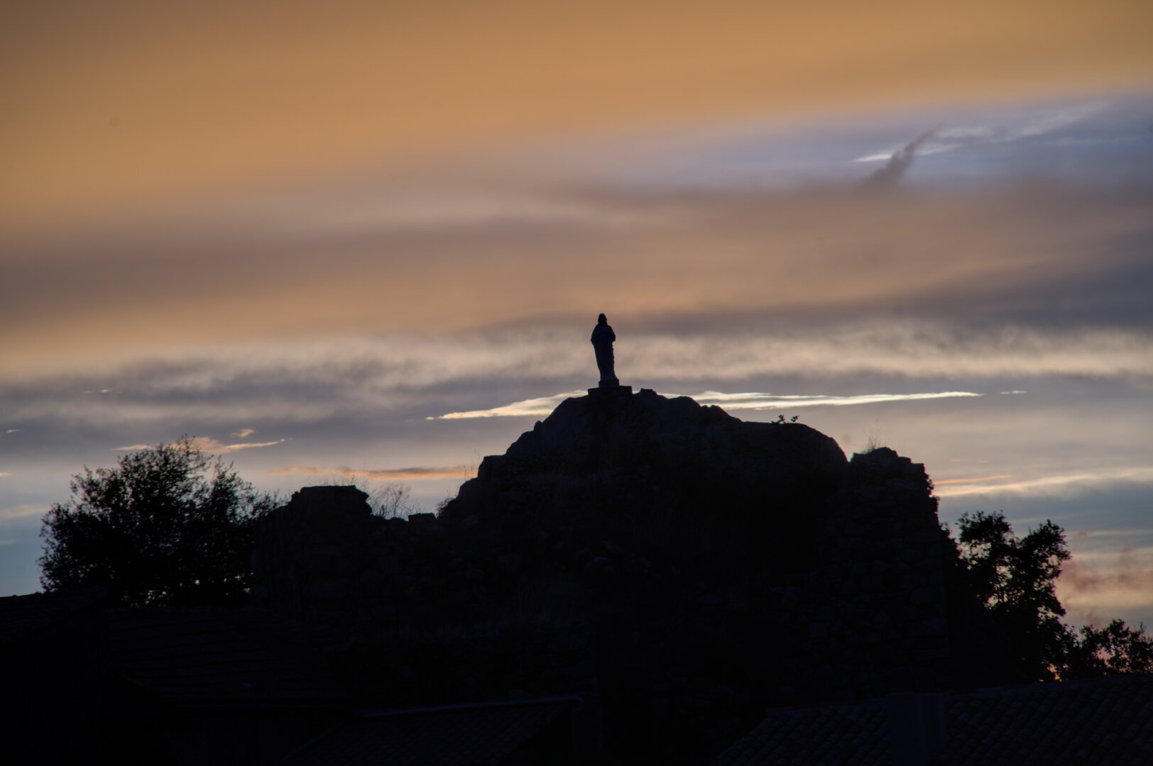 Une statue religieuse sur une butte rocheuse. La photo est prise au coucher du soleil, sur un ciel nuageux, il y a plein de couleurs dorées en arrière plan. Le premier plan avec la statue et la butte est sombre.