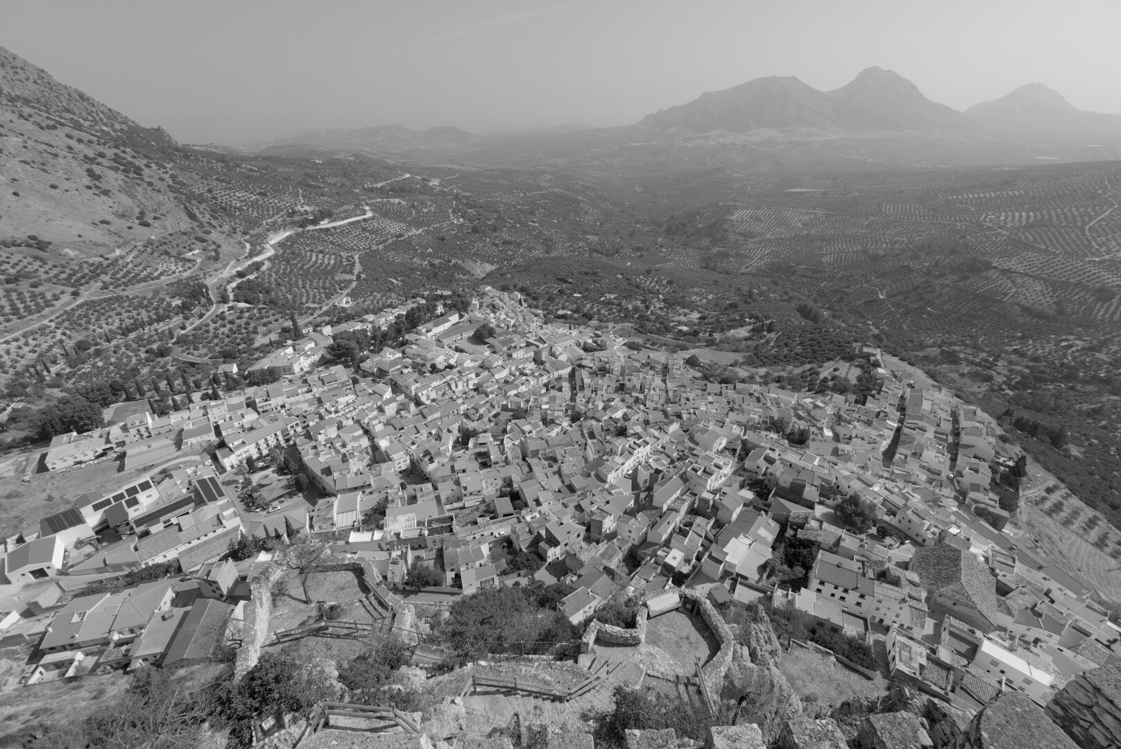 A black and white photo of a village, taken from above. The village is on the skirt of a mountain, there is another mountain to the left. Olive trees surround the village.