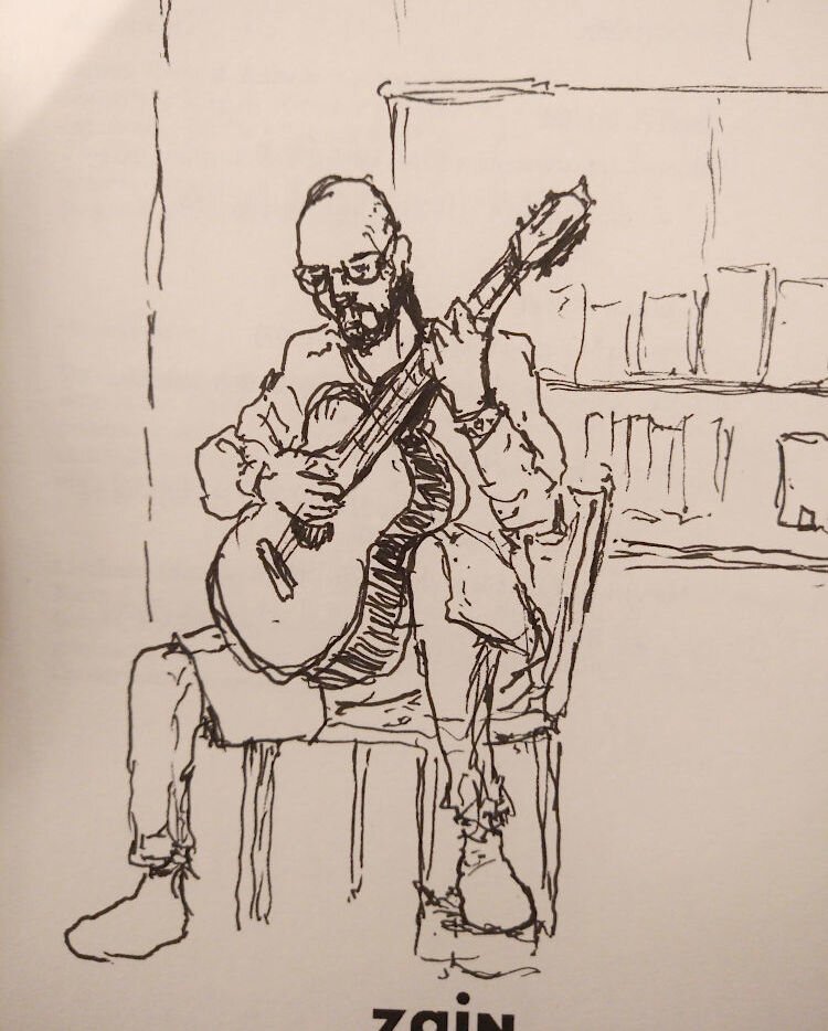 Pen drawing on a light brown paper, it's mostly made of lines, with little shading. In the image there's a man, he's slim, bald, with glasses and beard, probably in his thirties, playing an acoustic guitar, the instrument is placed diagonally, pointing up, with the bottom on his right leg and the middle part resting in his left leg, which is above a small support block on the ground to raise it higher. Behind him there's a few books, barely visible, drawn with a few lines. In the bottom of the page there's the uppermost part of the printed text 