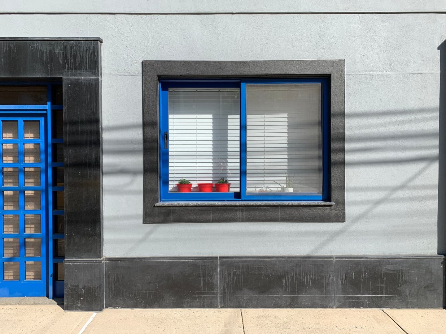 A window of a grey building with bright blue panes and a thick black border has three well placed red plant pots.