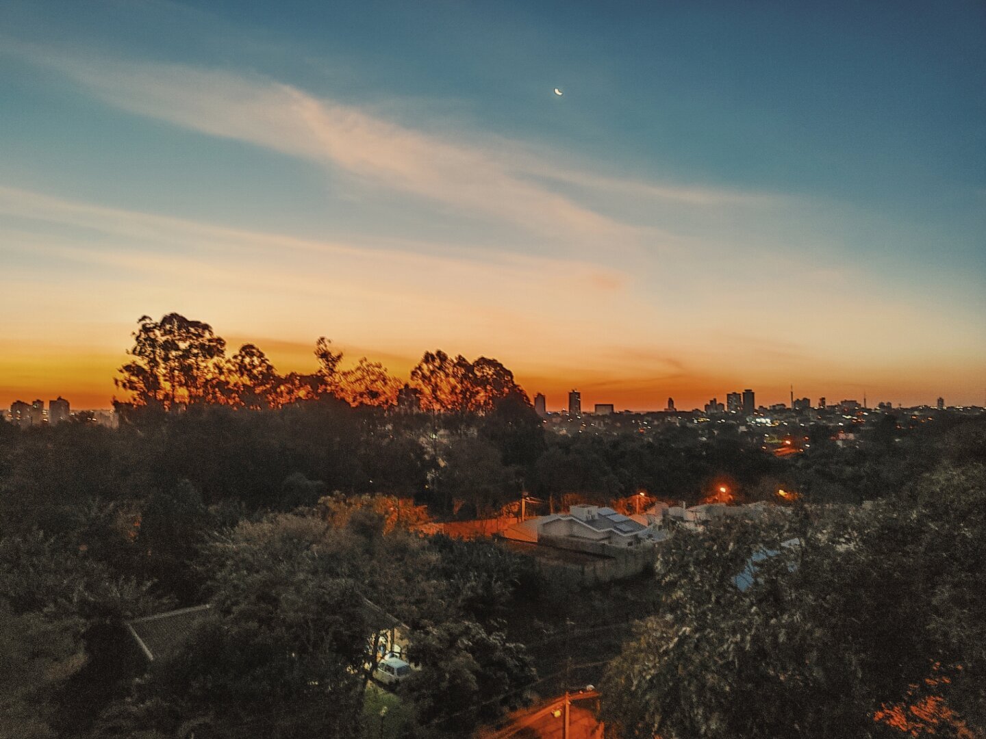 Retrato de paisagem da cidade, com algumas casas e muitas árvores. Ao fundo, o pôr do sol em tons amarelos e laranjas e o céu ainda azul. Também aparece a lua em fase crescente.