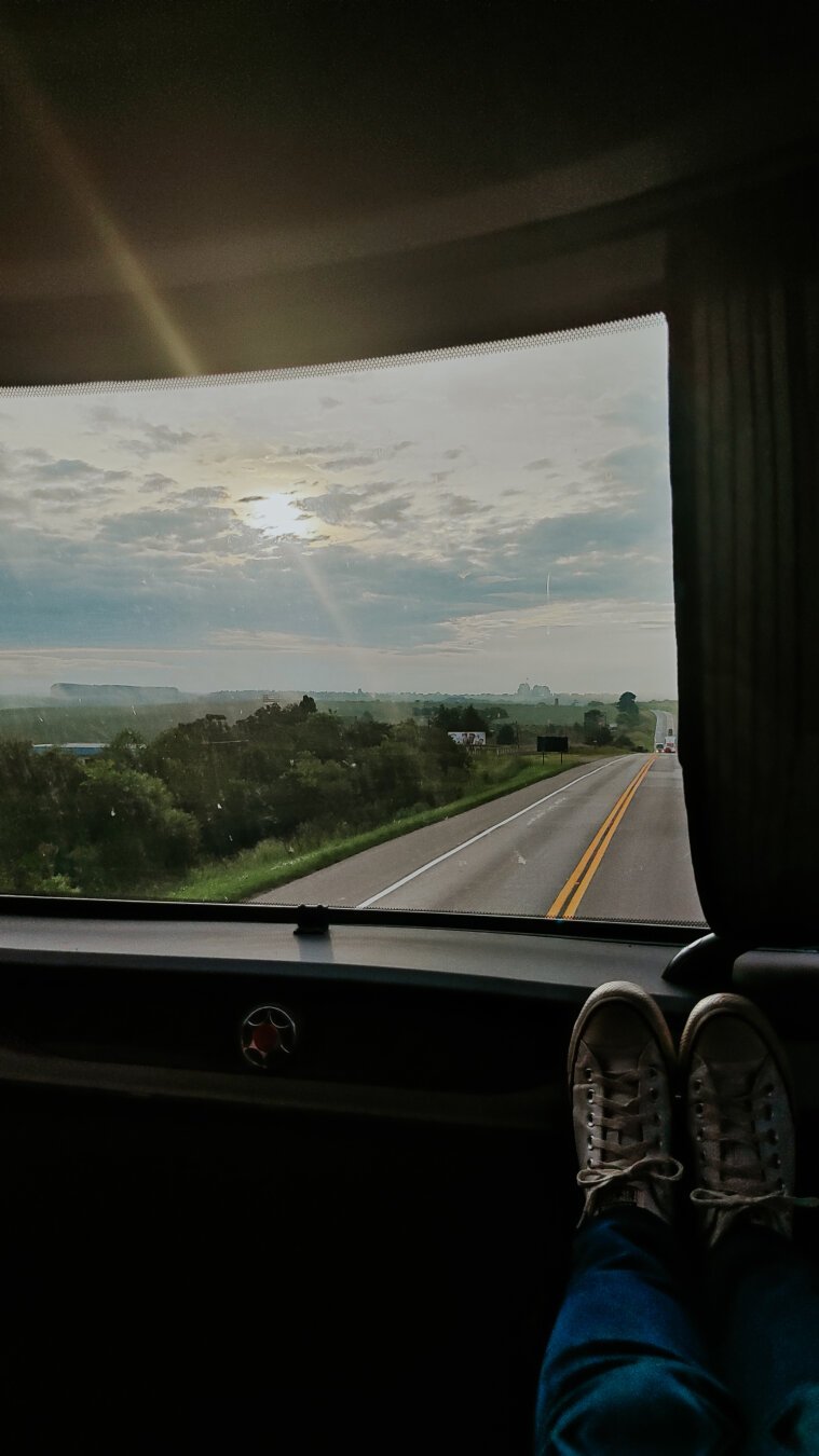 Retrato tirado dentro de um ônibus de viagem, mostra uma janela e nela se vê uma estrada, algumas árvores, um campo verde, o céu com algumas nuvens e sol nascendo entre elas. Dentro do ônibus, ao redor da janela, está escuro e aparece um par de tênis branco um pouco abaixo da janela.