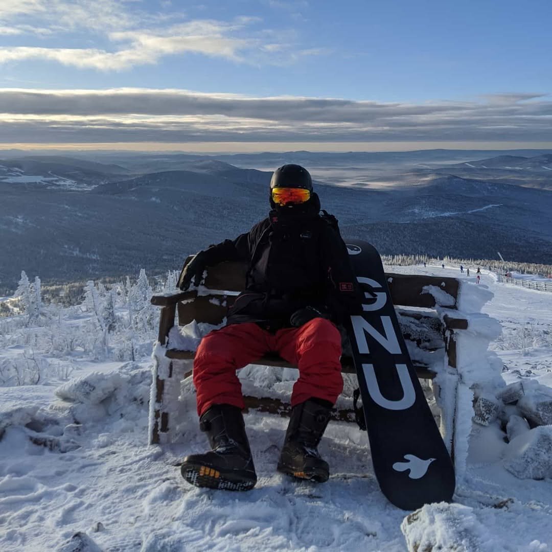 A man in a snoboarding equipment sitting on the frozen bench on the top of the hill, forests, snow and mountains are around. The snowboard with big G N U letters on the bottom.