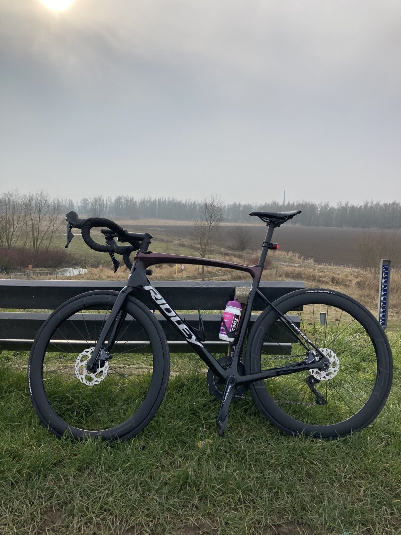 Black and red race bike standing against a bench in the outside. View of grey sky and a tiny sun and in between the riverside landscape