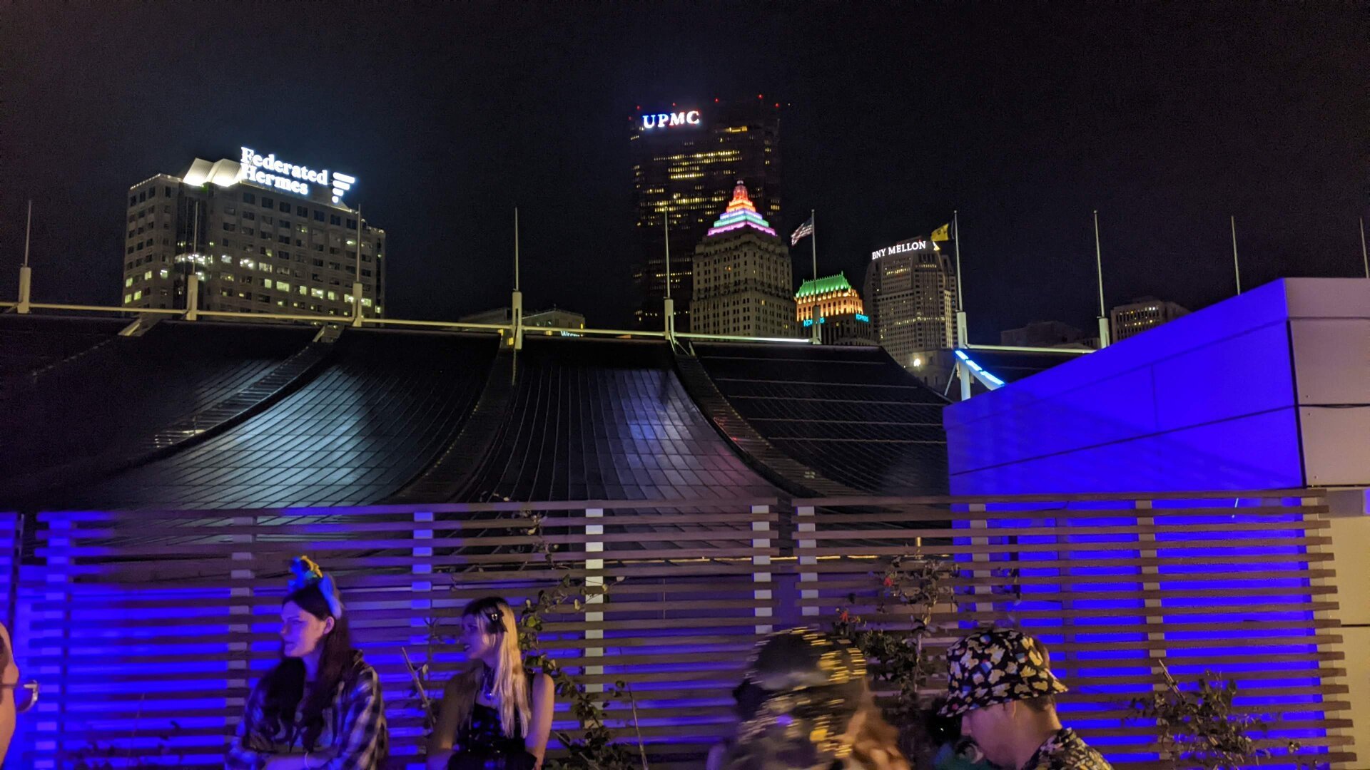 The downtown Pittsburgh skyline as viewed from the roof of the convention center. The roof slopes upward and is slightly illuminated in purple light. Behind it, are four of the tallest buildings in Pittsburgh with their lights on. Flags fly in the distance on flag polls, including an Anthrocon flag.