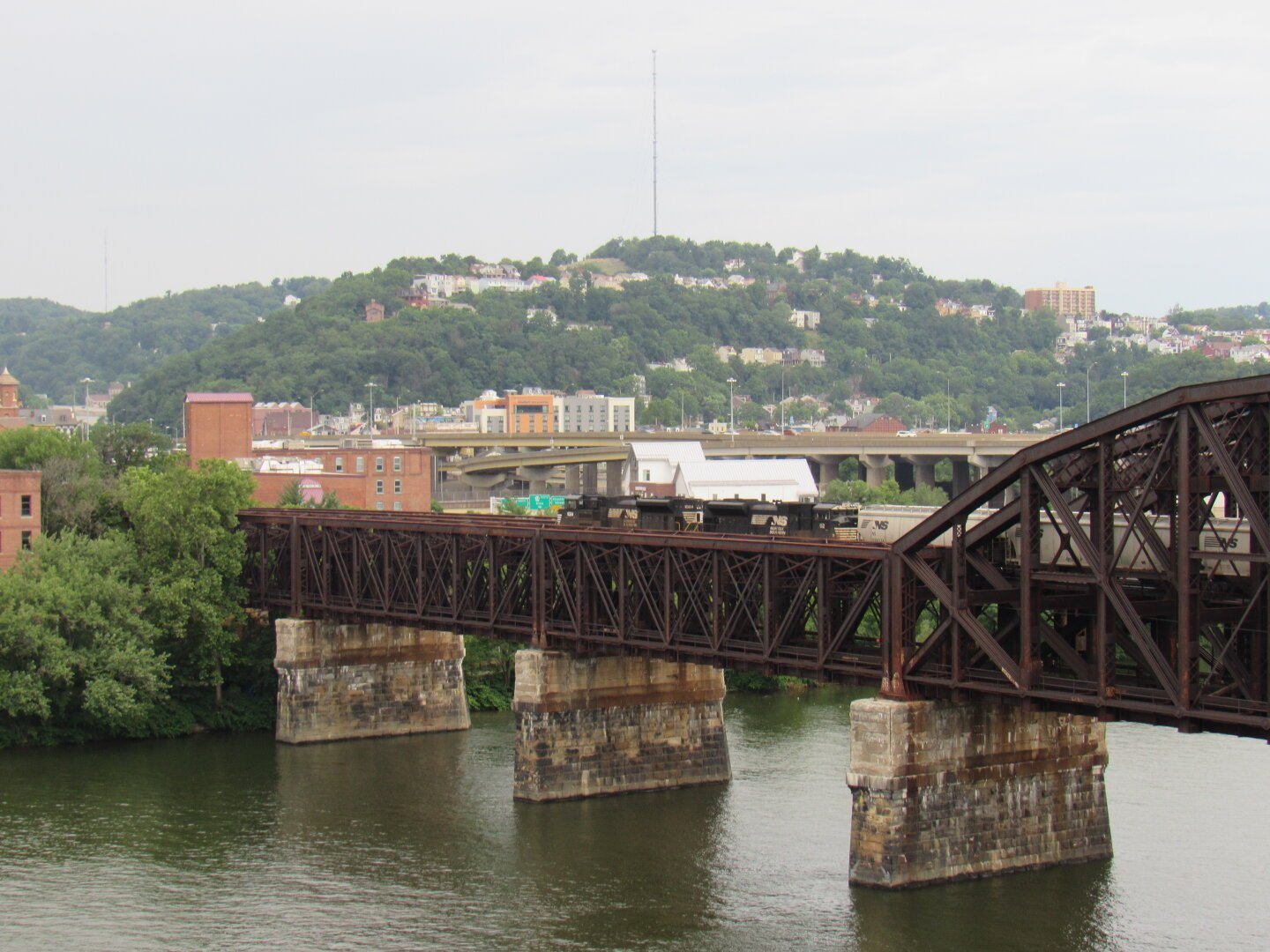 A freight train crossing a dedicated freight bridge in Pittsburgh, PA. Behind the bridge is an overpass with cars. Further in the distance is a large, tree-covered hill dotted with many buildings and homes.