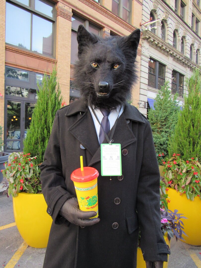 A black werewolf fursuiter with yellow eyes wears a suit and tie with a peacoat. He has on black gloves to complete the outfit. In his hand is a lemonade.