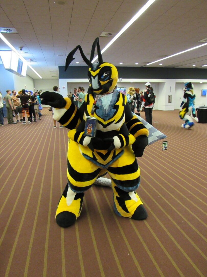 A honey bee fursuiter named Rampage at Anthrocon 2024. Their suit has four arms and two legs. They also have a smaller conbadge on their wing that says 