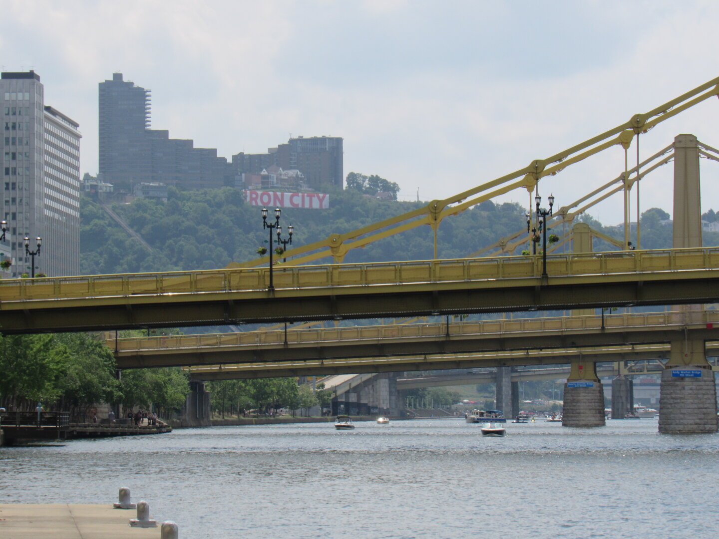 A shot of a yellow bridge and the Allegheny River below it. Behind that bridge are another three bridges. In the distance is small, tree-covered mountain. An apartment complex sits at the top of the small mountain and a banner that reads 