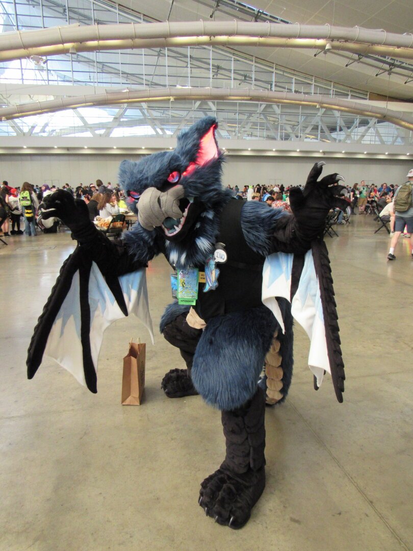 A Nargacua from the Monster Hunter games fursuiter named Inq. His eyes and ears glow red, and he has a dapper black vest on.