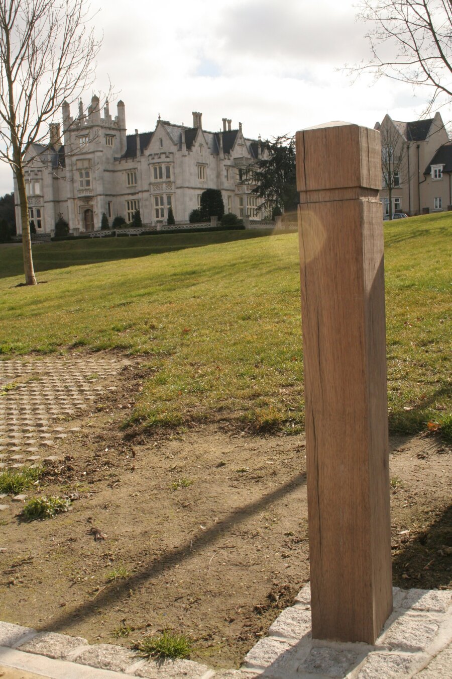 A woodwn post is the subject of this photo. in the background is a large old white building and a park green in the mid-ground.