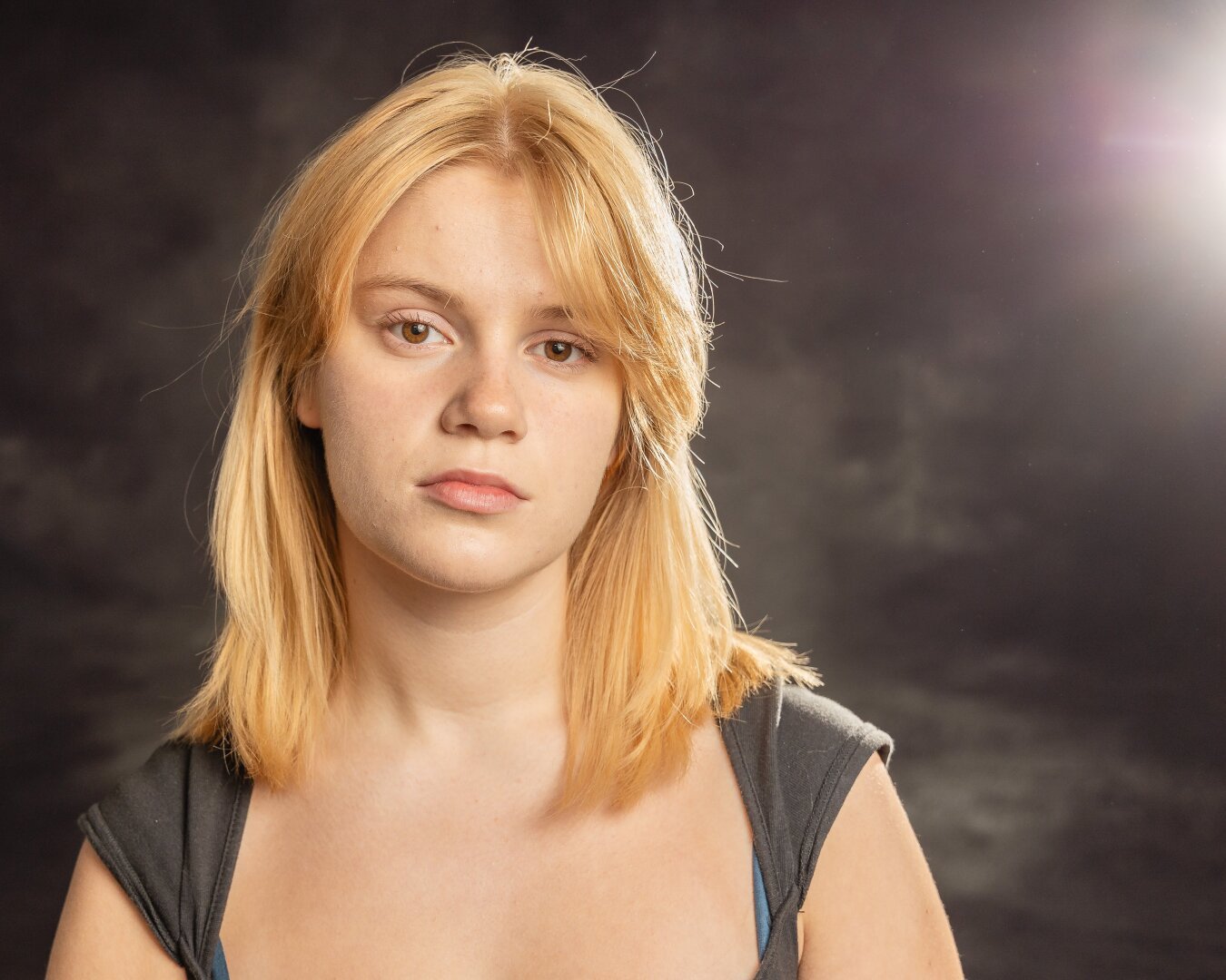 portrait of a young women with long  blond hair. A light to camera right highlights her hair. the background is a mottled dark grey.