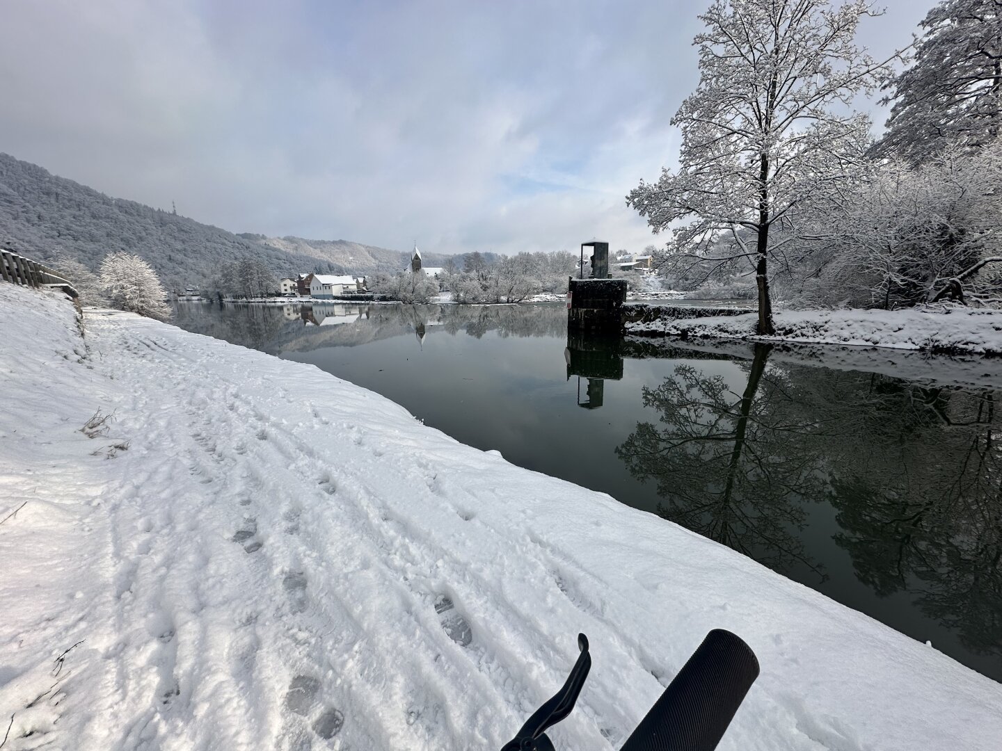 Snow-covered riverbank with footprints, trees laden with snow, calm river reflecting trees and buildings, partial view of a bicycle's handlebar in the foreground.